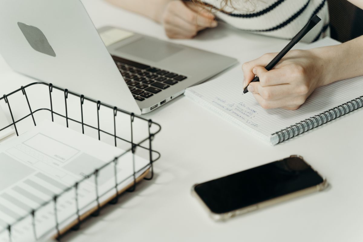 A person writing notes in a notebook beside an open laptop.